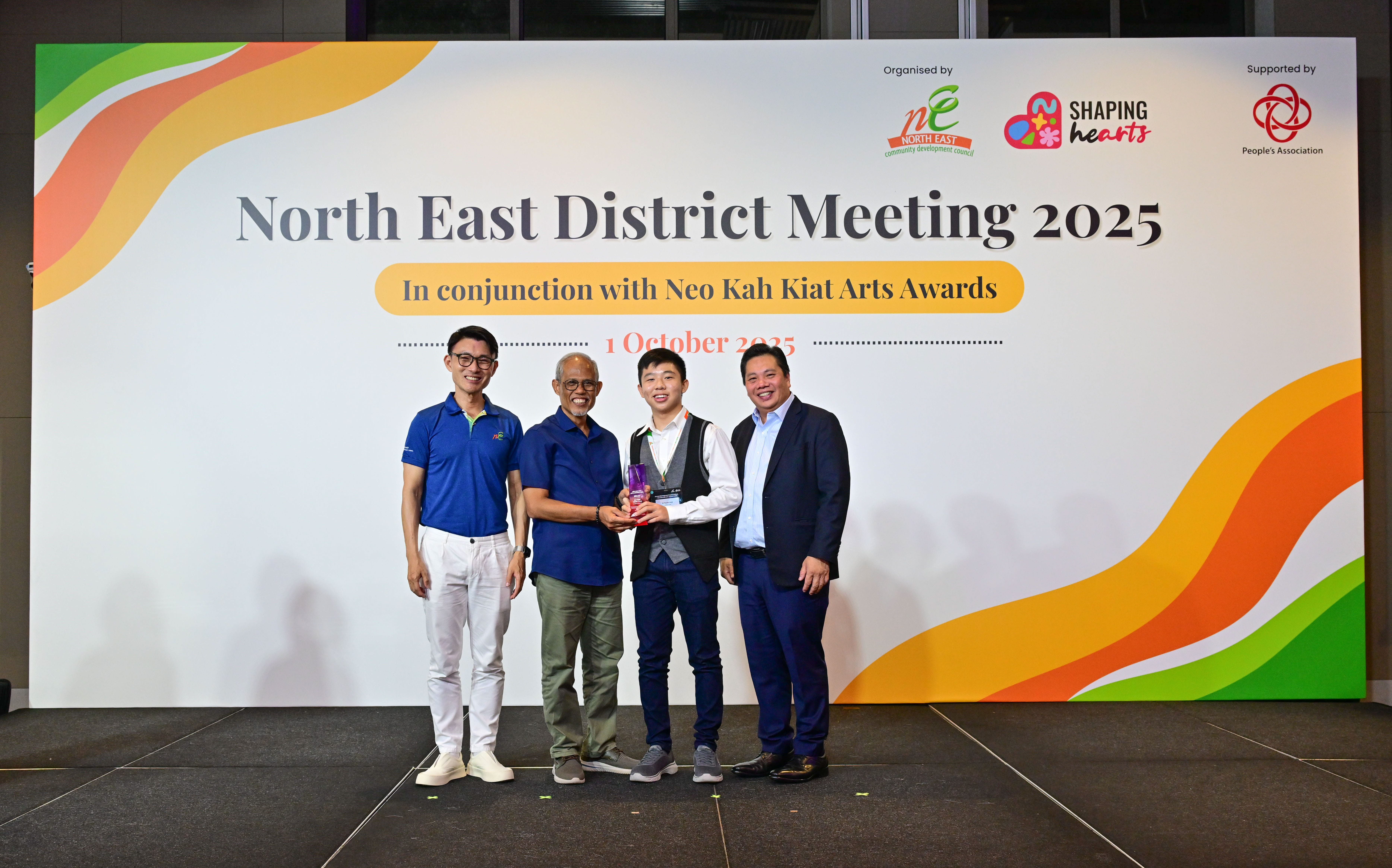 Award recipient standing on stage with the presenter, holding a acrylic plaque and posing for a group photo during the award ceremony, with a backdrop reading ‘North East District Meeting 2025 In conjunction with Neo Kah Kiat Arts Awards'